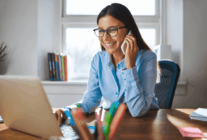 A young women smiling speaks on the phone while looking at her laptop.
