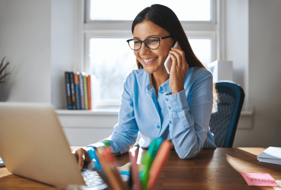 A young women smiling speaks on the phone while looking at her laptop.