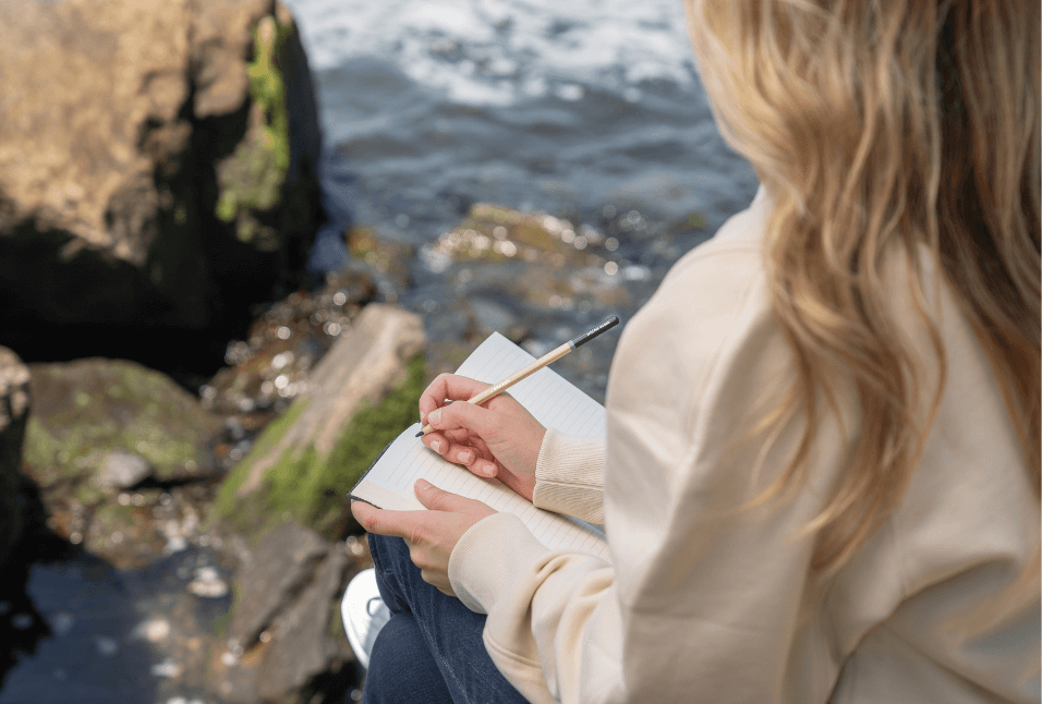 Woman sitting on rocks by the water writing in a journal.